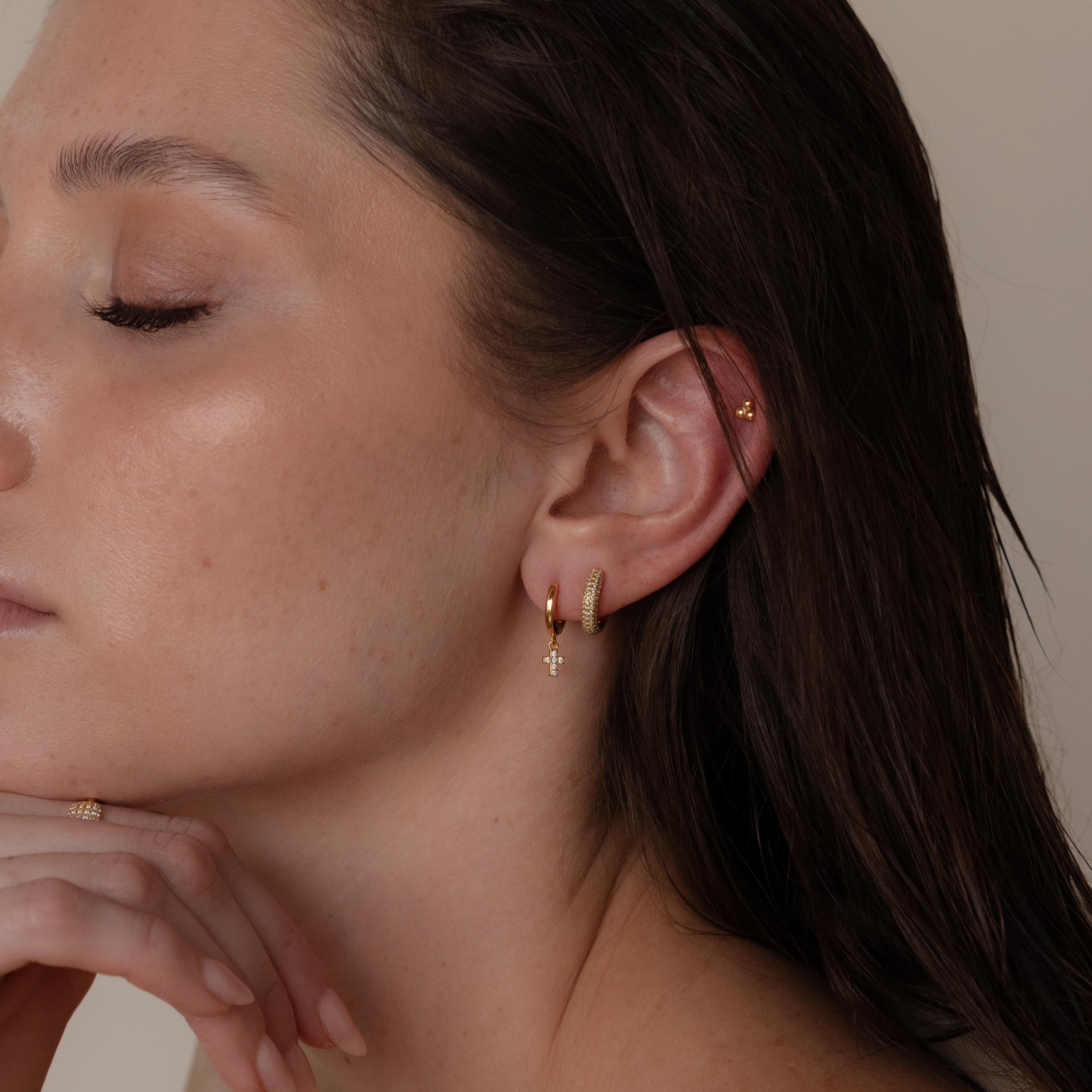 With her eyes closed, a woman wears the Pave Cross Huggies and a ring, gently resting her chin on her hand against a neutral background.