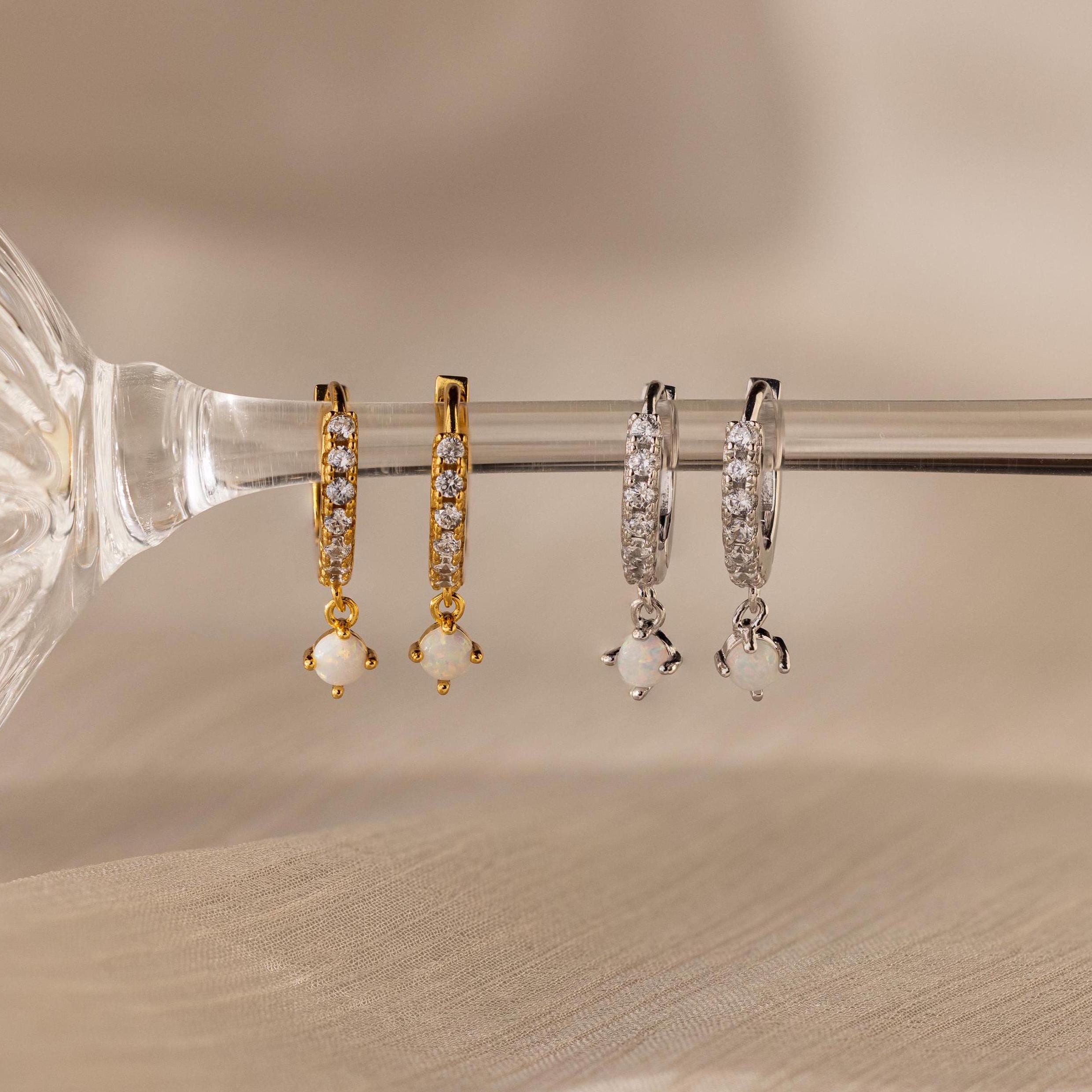 Two pairs of crystal-studded hoop earrings, including the gold Pavé Opal Drop Huggies and classic silver diamond earrings, are displayed on a glass rod.