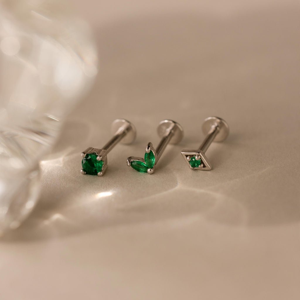 The Emerald Flat Back Earring Set features three silver screw-back earrings with green stones, elegantly displayed on a beige surface under soft lighting.