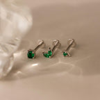 The Emerald Flat Back Earring Set features three silver screw-back earrings with green stones, elegantly displayed on a beige surface under soft lighting.
