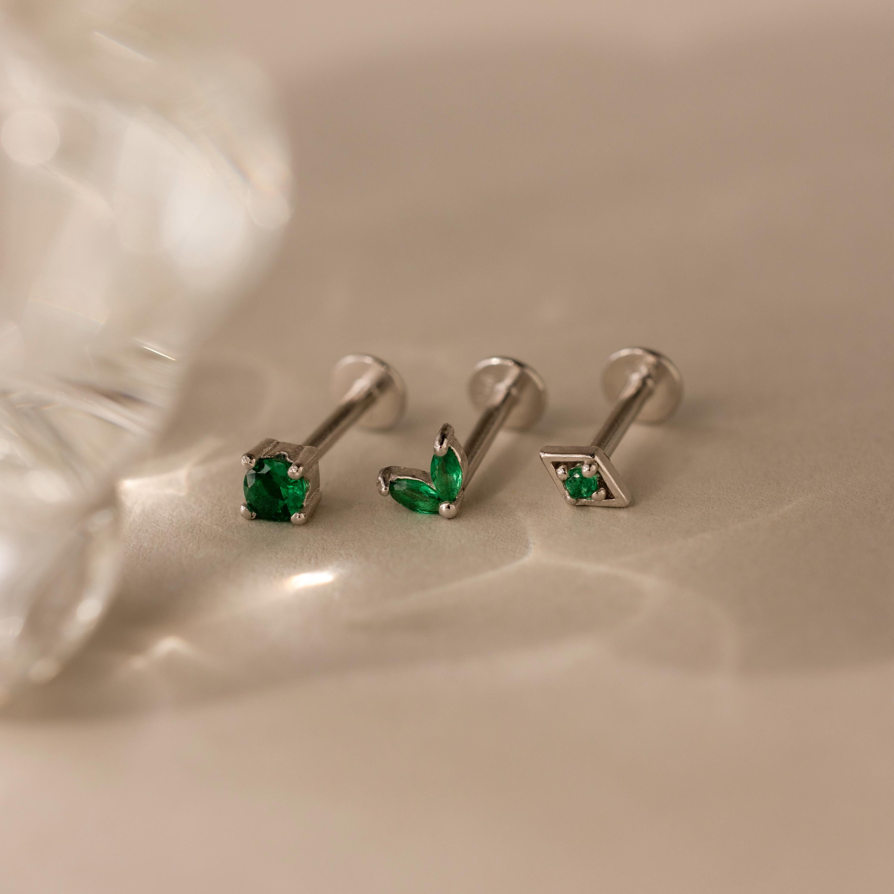 The Emerald Flat Back Earring Set features three silver screw-back earrings with green stones, elegantly displayed on a beige surface under soft lighting.