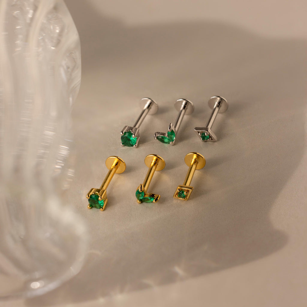 The Emerald Flat Back Earring Set features six stud earrings—three silver and three gold—each adorned with a small green gemstone, ideal as emerald or birthstone studs, all presented on a beige surface.