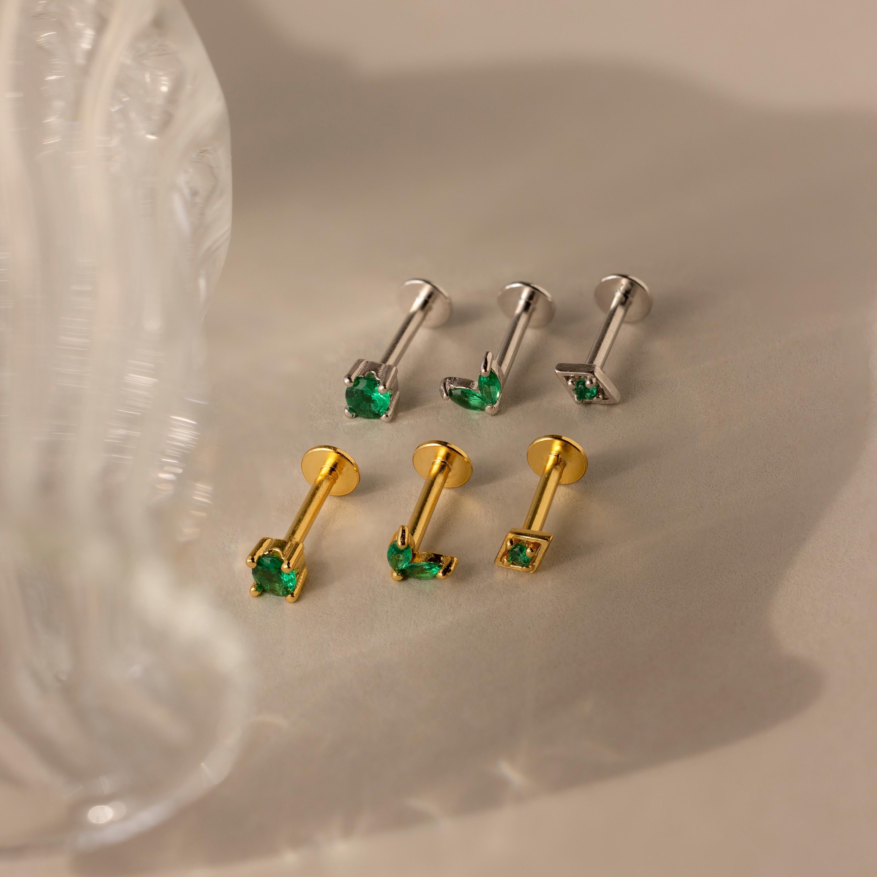 The Emerald Flat Back Earring Set features six stud earrings—three silver and three gold—each adorned with a small green gemstone, ideal as emerald or birthstone studs, all presented on a beige surface.