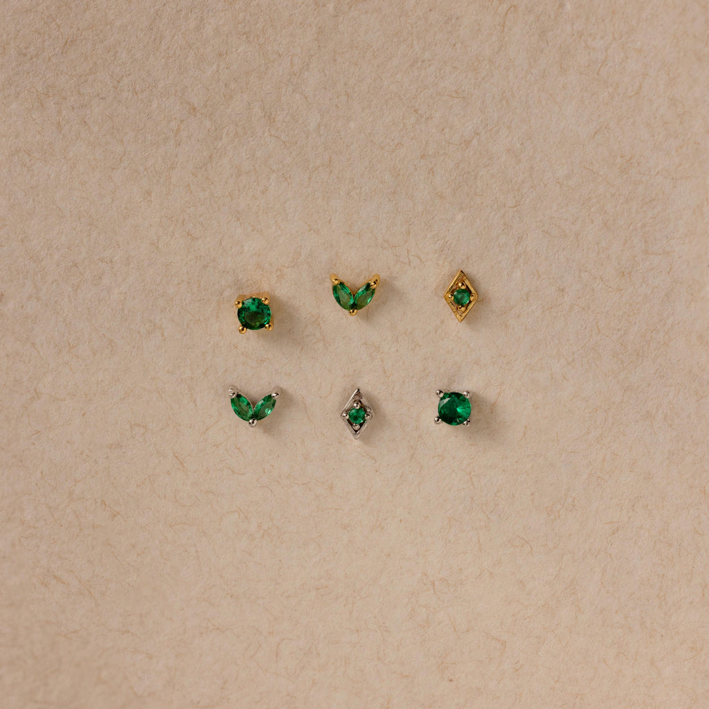 The Emerald Flat Back Earring Set features six small emerald studs in gold and silver settings, displayed in two rows on a beige textured background—ideal as birthstone earrings.