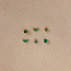 The Emerald Flat Back Earring Set features six small emerald studs in gold and silver settings, displayed in two rows on a beige textured background—ideal as birthstone earrings.