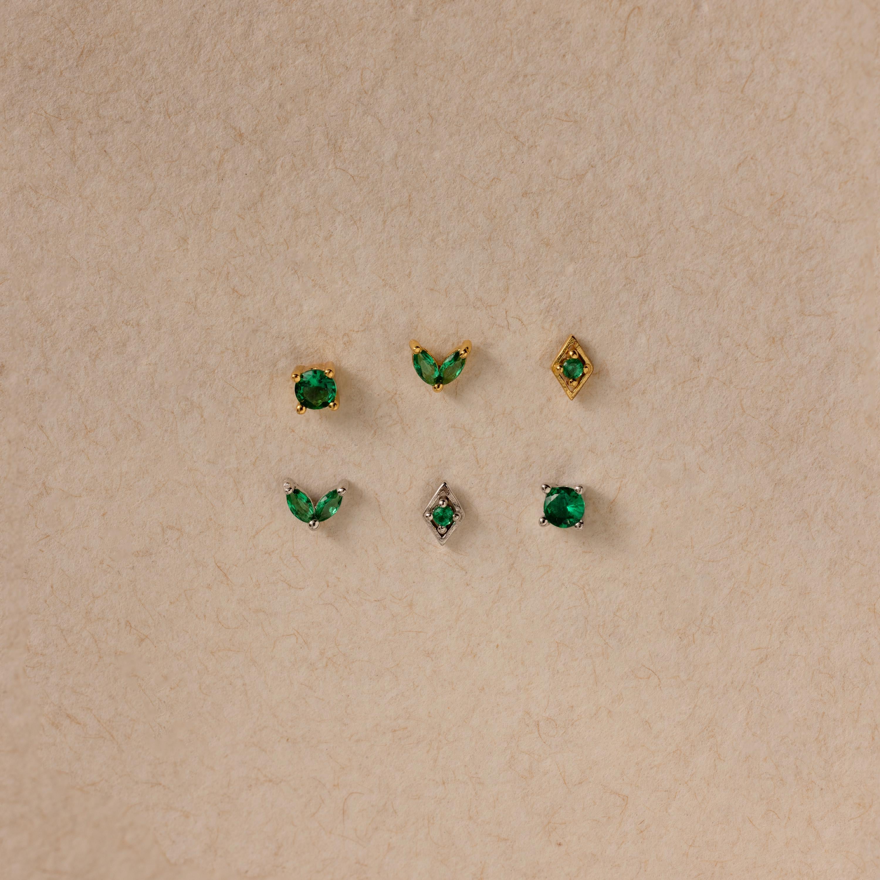 The Emerald Flat Back Earring Set features six small emerald studs in gold and silver settings, displayed in two rows on a beige textured background—ideal as birthstone earrings.