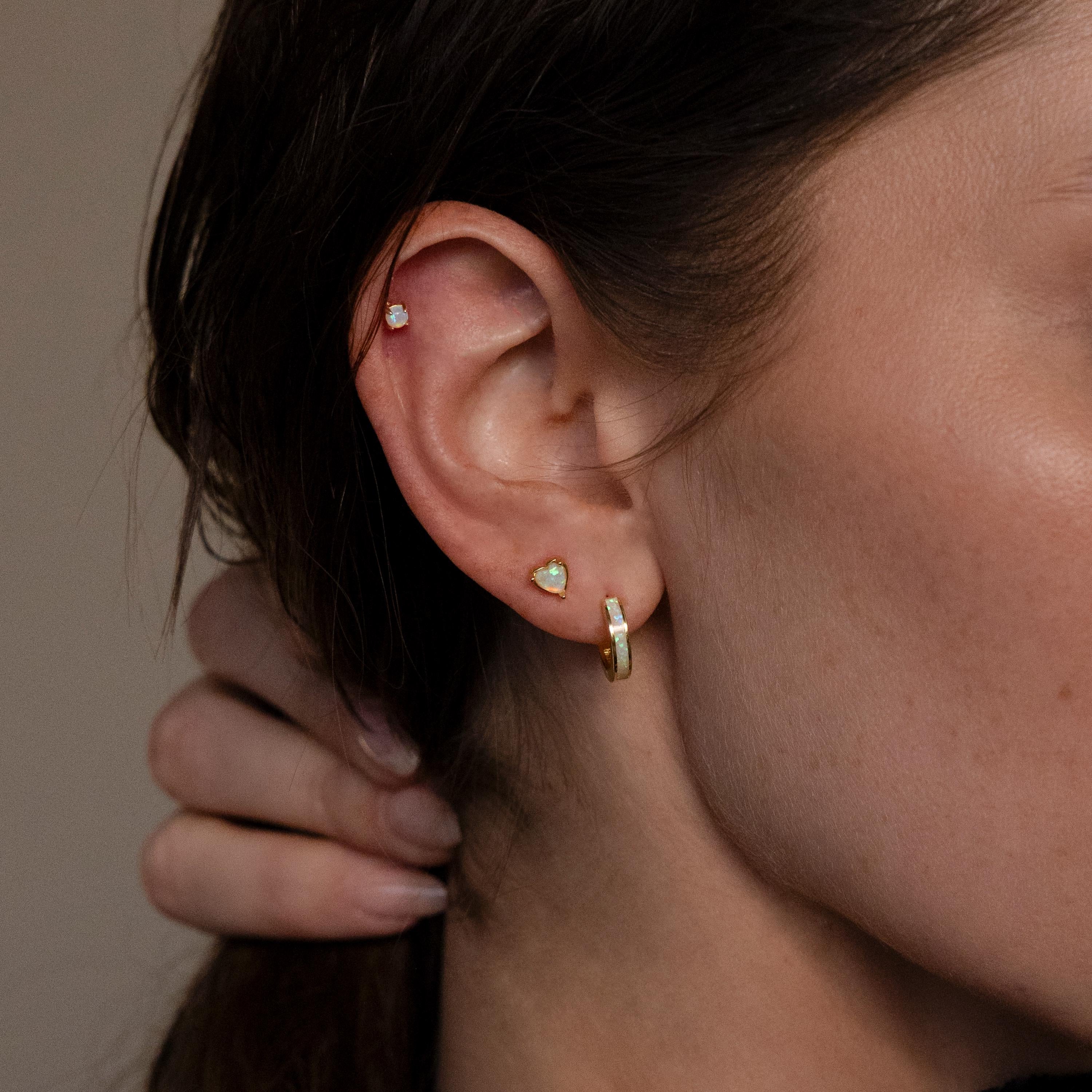 Close-up of a person's ear with three gold earrings, including Opal Heart Flat Back Studs, and their hand resting on the back of their neck.