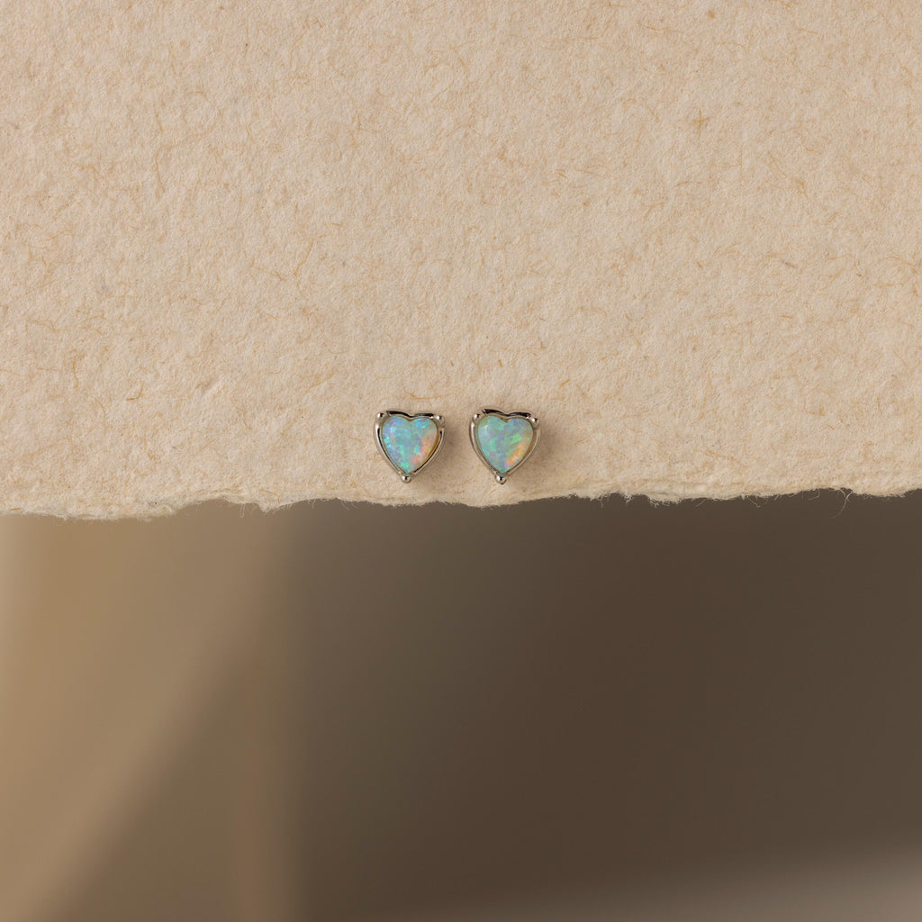 The Opal Heart Flat Back Studs are shown on a textured beige surface, offering a delicate addition to your gemstone earrings collection.
