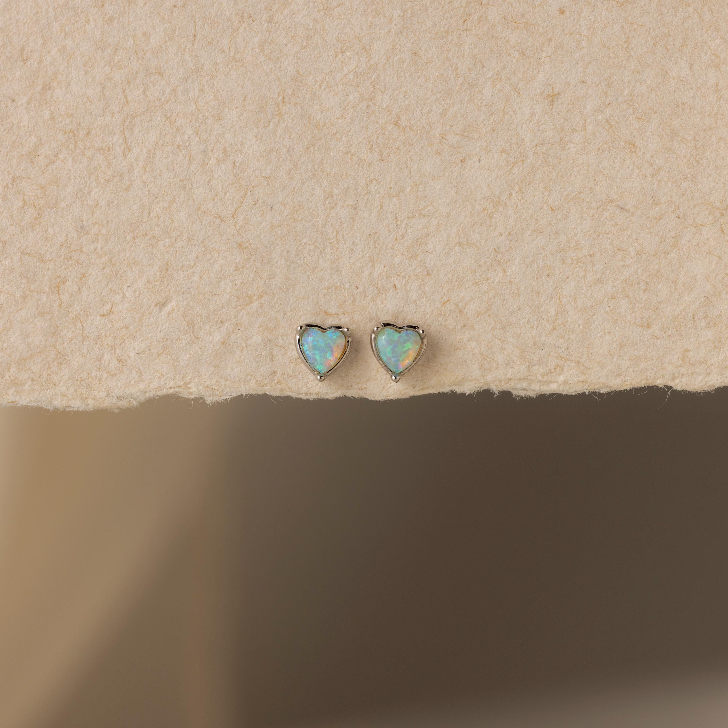 The Opal Heart Flat Back Studs are shown on a textured beige surface, offering a delicate addition to your gemstone earrings collection.
