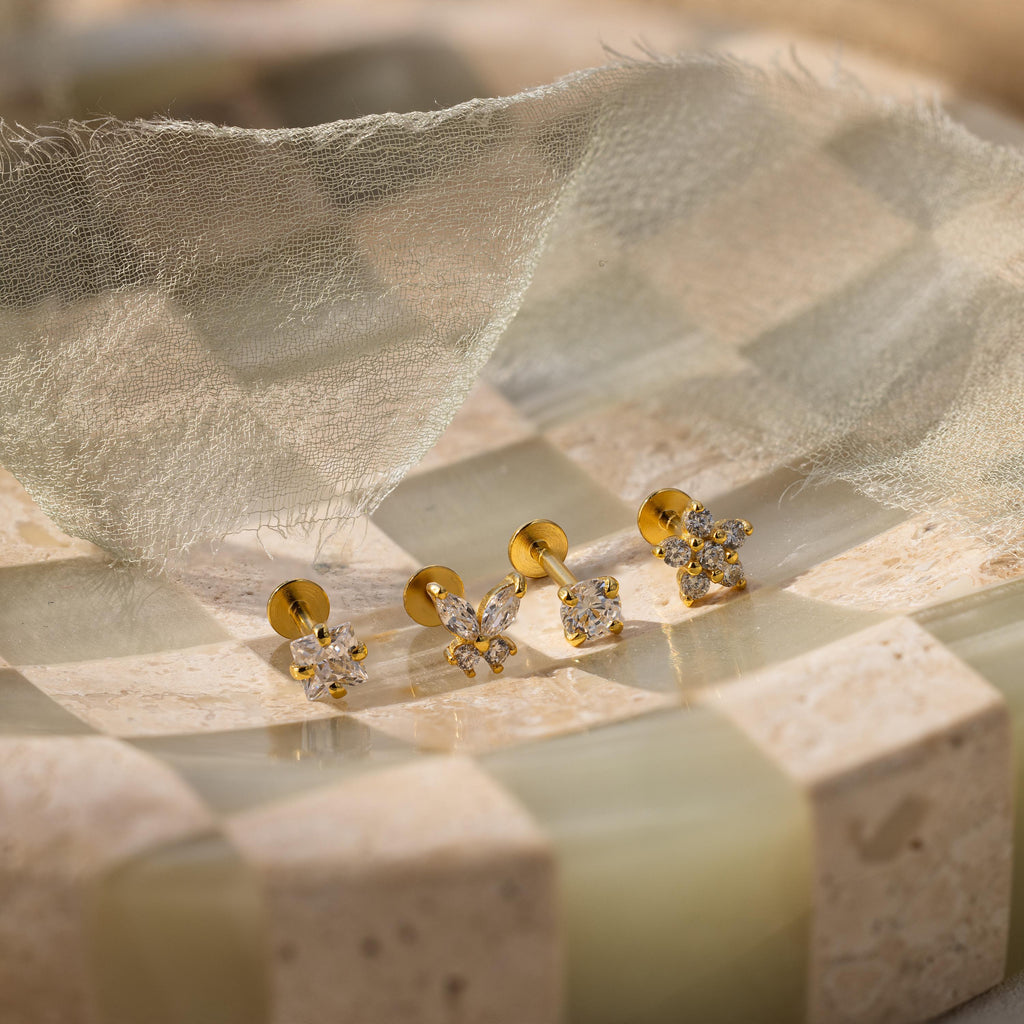 The Pave Assortment Flat Back Set features three pairs of gold and pavé diamond flat-back studs displayed on a checkered stone surface with sheer fabric above for an elegant presentation.
