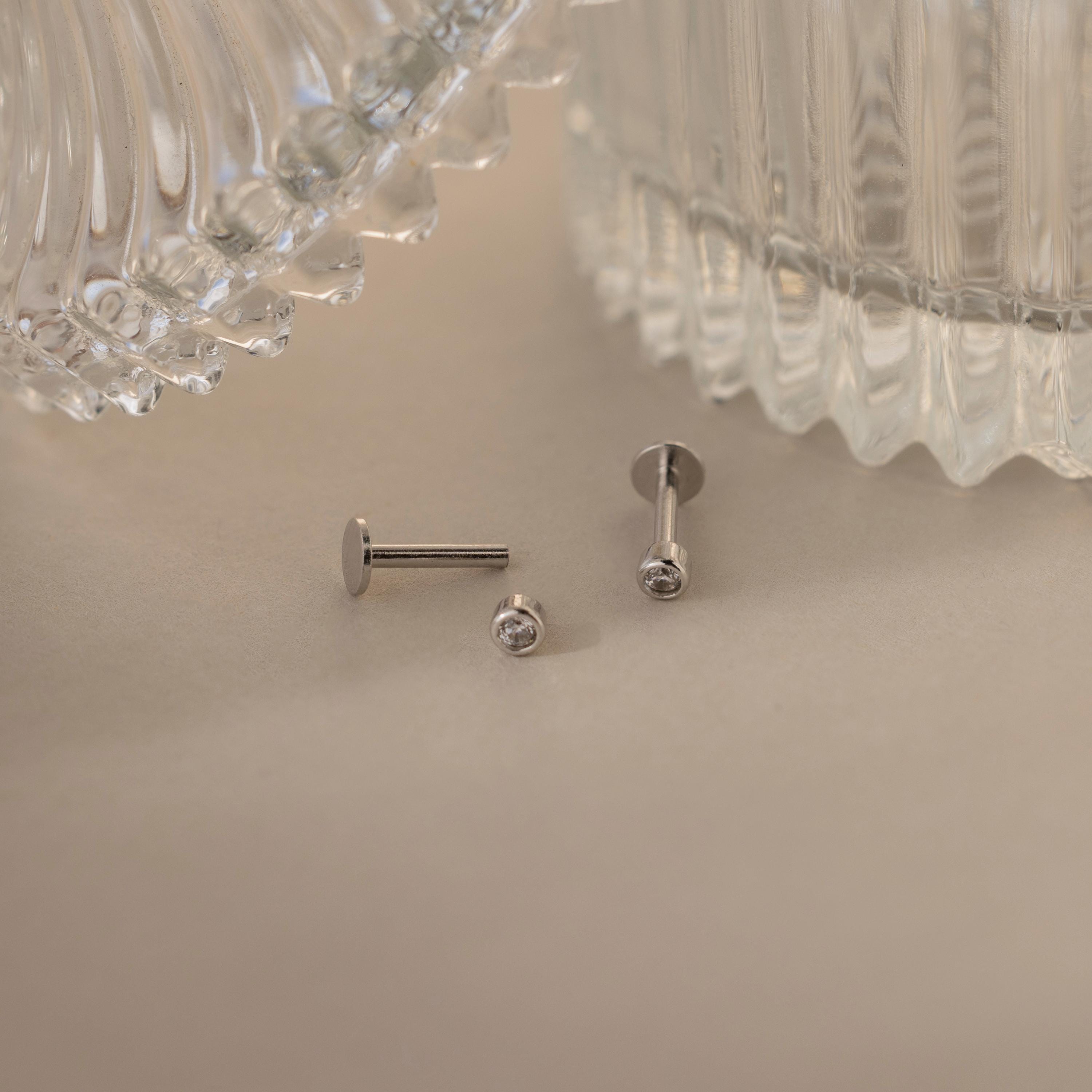 Two Small Diamond Bezel Flatback Studs with clear gems rest on a beige surface near ribbed glass containers.