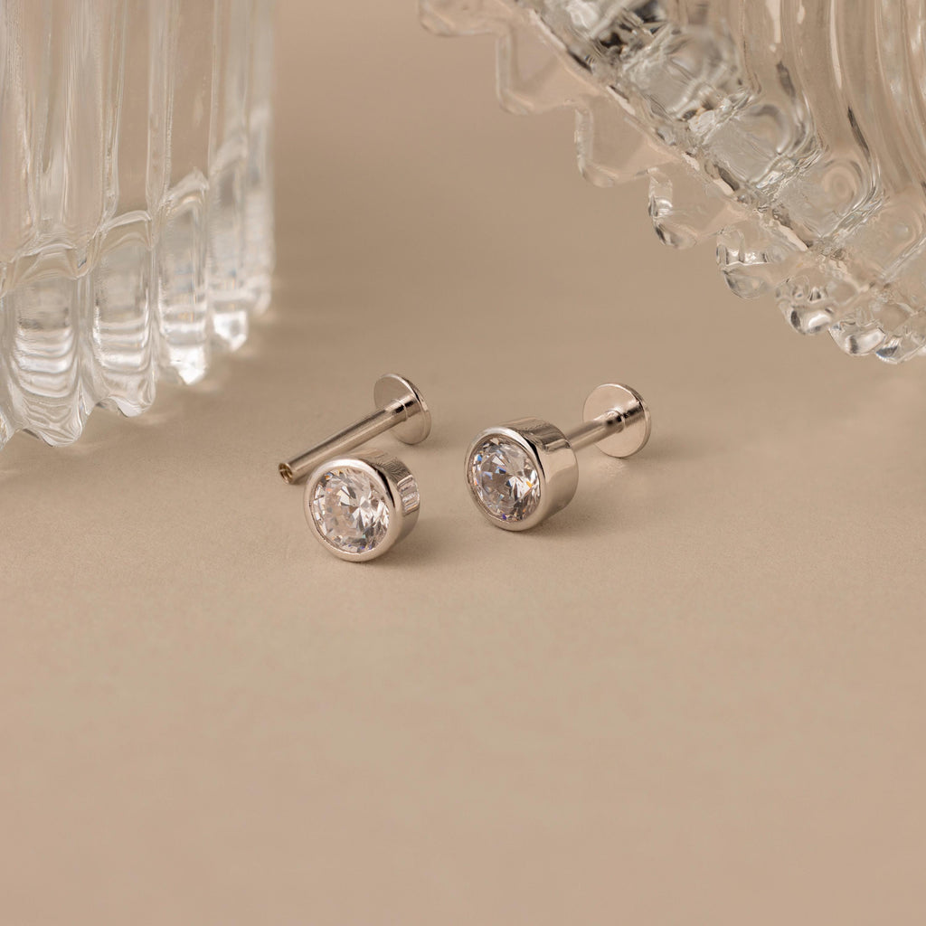 Large Diamond Bezel Flatback Studs featuring clear gemstones are displayed on a beige surface near glass objects.