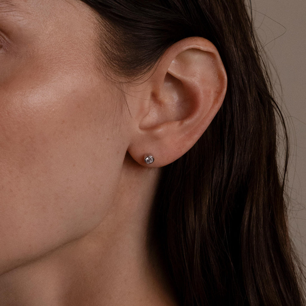 Close-up of a woman's ear with straight brown hair tucked behind it, showcasing the Medium Diamond Bezel Flatback Studs—an elegant addition to her jewelry collection.