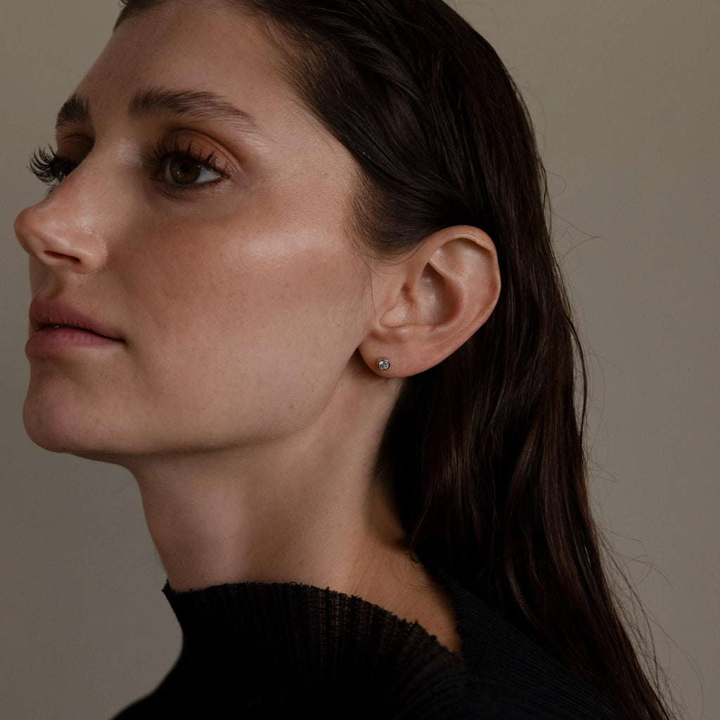 Woman with wet, dark hair and a black sweater wears Medium Diamond Bezel Flatback Studs from her jewelry collection while looking to the left.