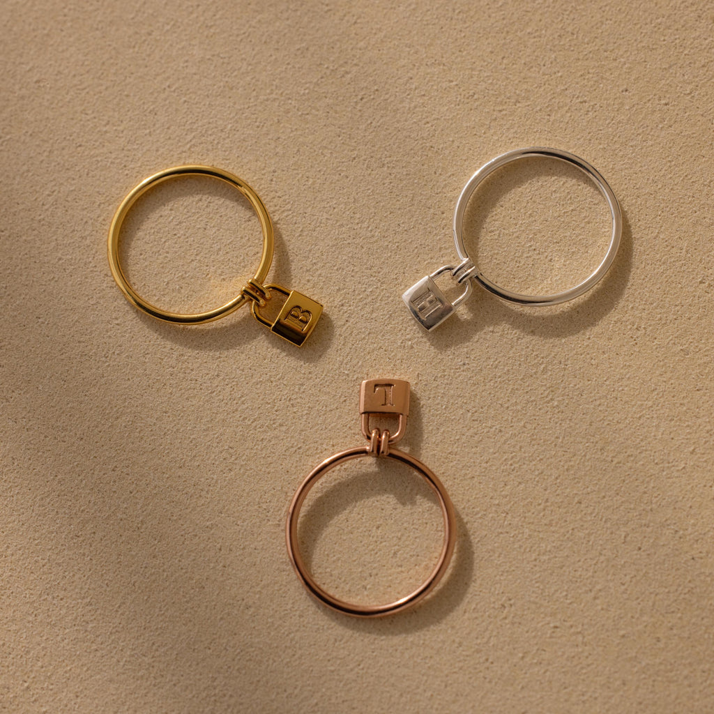The Initial Padlock Drop Ring features three metal bands with small padlock charms in gold, silver, and rose gold—ideal as an initial ring or for adding a personalized touch to your jewelry collection.