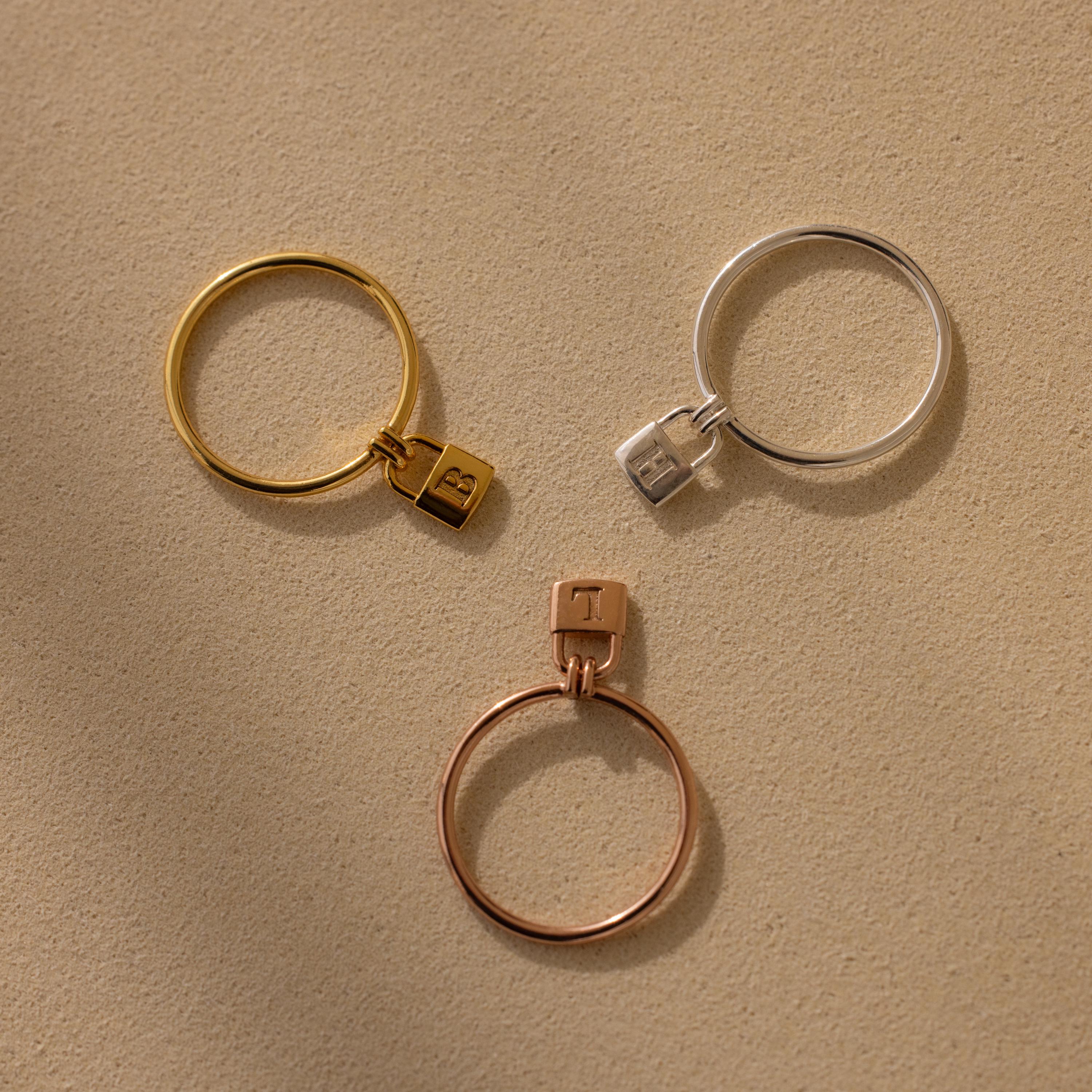 The Initial Padlock Drop Ring features three metal bands with small padlock charms in gold, silver, and rose gold—ideal as an initial ring or for adding a personalized touch to your jewelry collection.