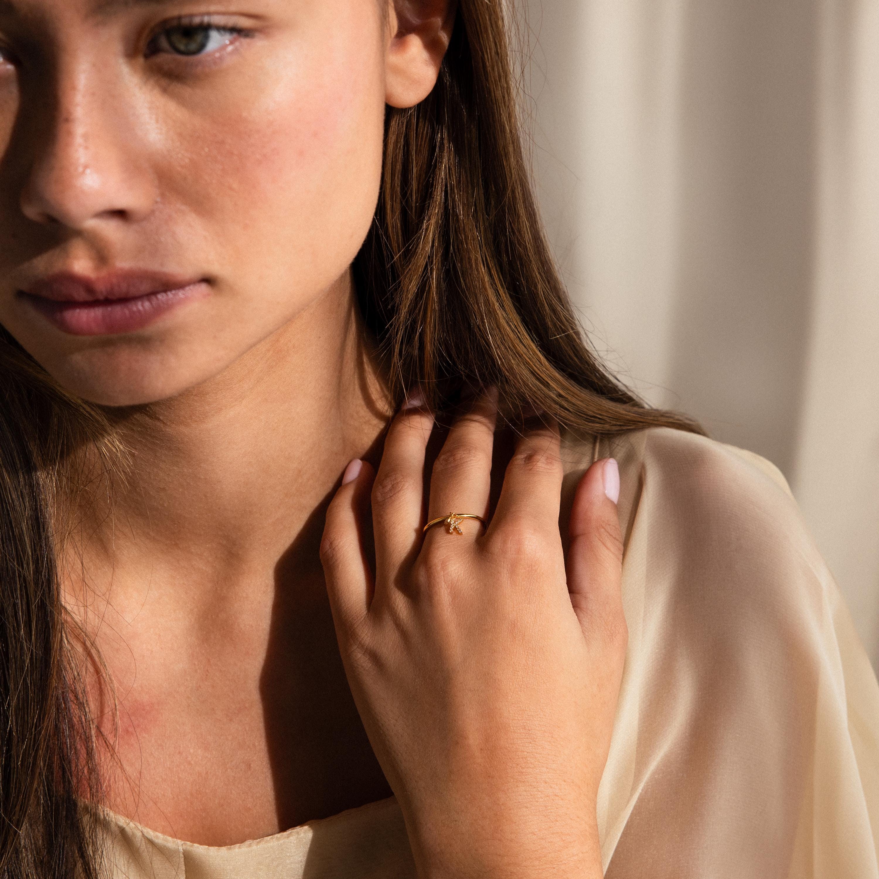 Woman with long brown hair, dressed in a cream blouse, touches her chest while wearing a gold Pave Initial Birthstone Drop Ring.