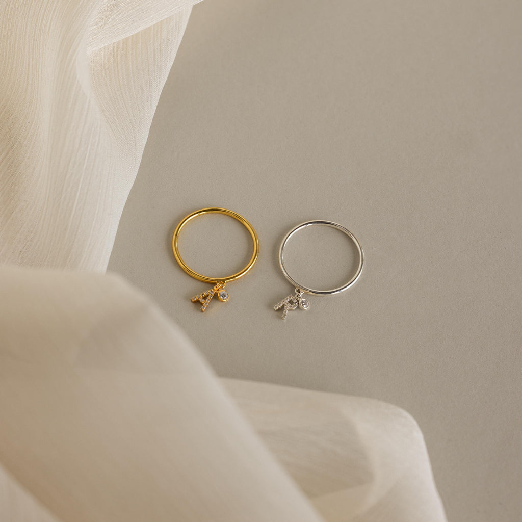 The Pave Initial Birthstone Drop Ring, featuring sparkling charms, rests on a neutral surface with sheer fabric draped nearby.