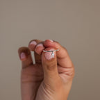 A hand holds the Pave Initial Birthstone Drop Ring, featuring a delicate silver band and a small dangling accent, ideal for personalizing with your birthstone, shown against a plain background.
