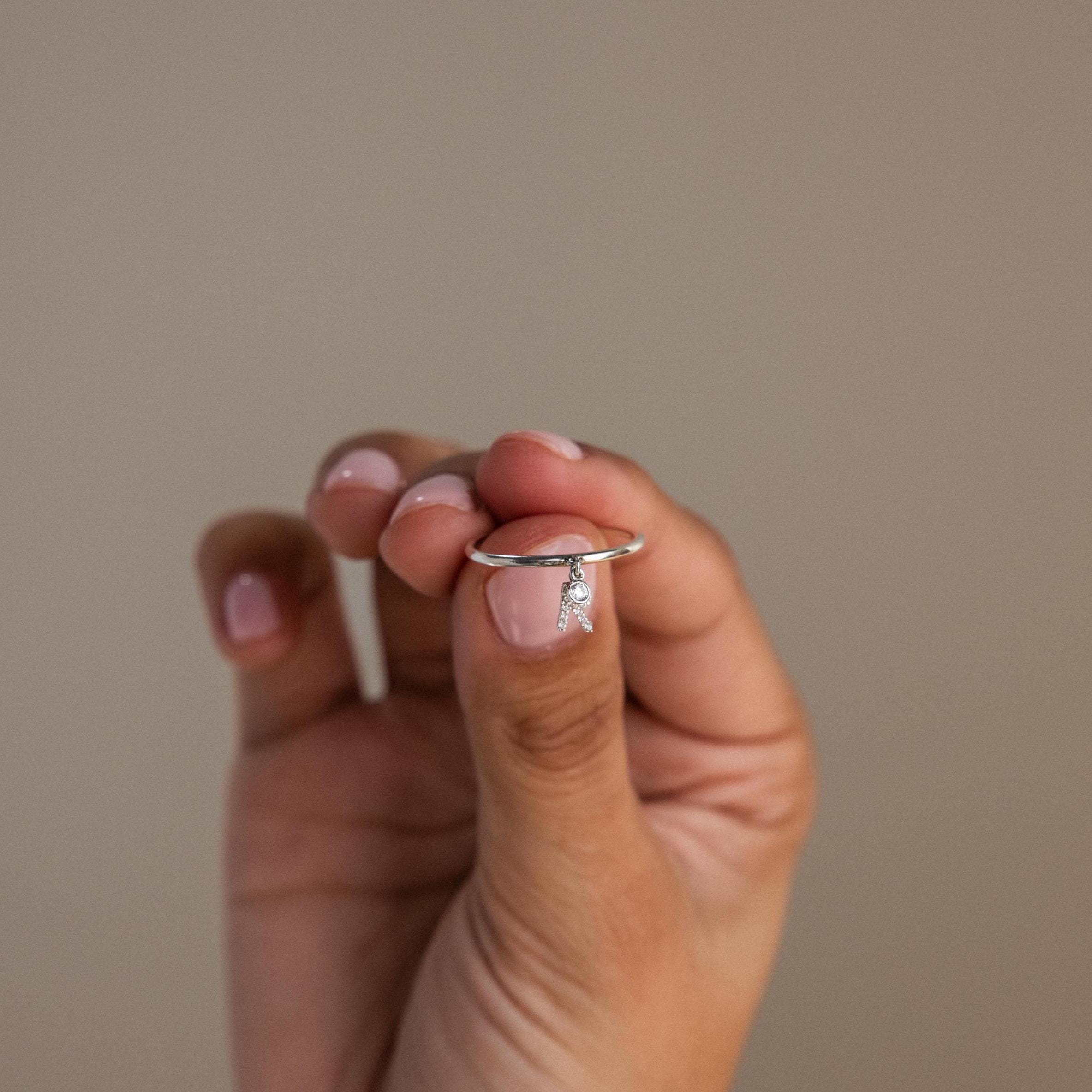 A hand holds the Pave Initial Birthstone Drop Ring, featuring a delicate silver band and a small dangling accent, ideal for personalizing with your birthstone, shown against a plain background.