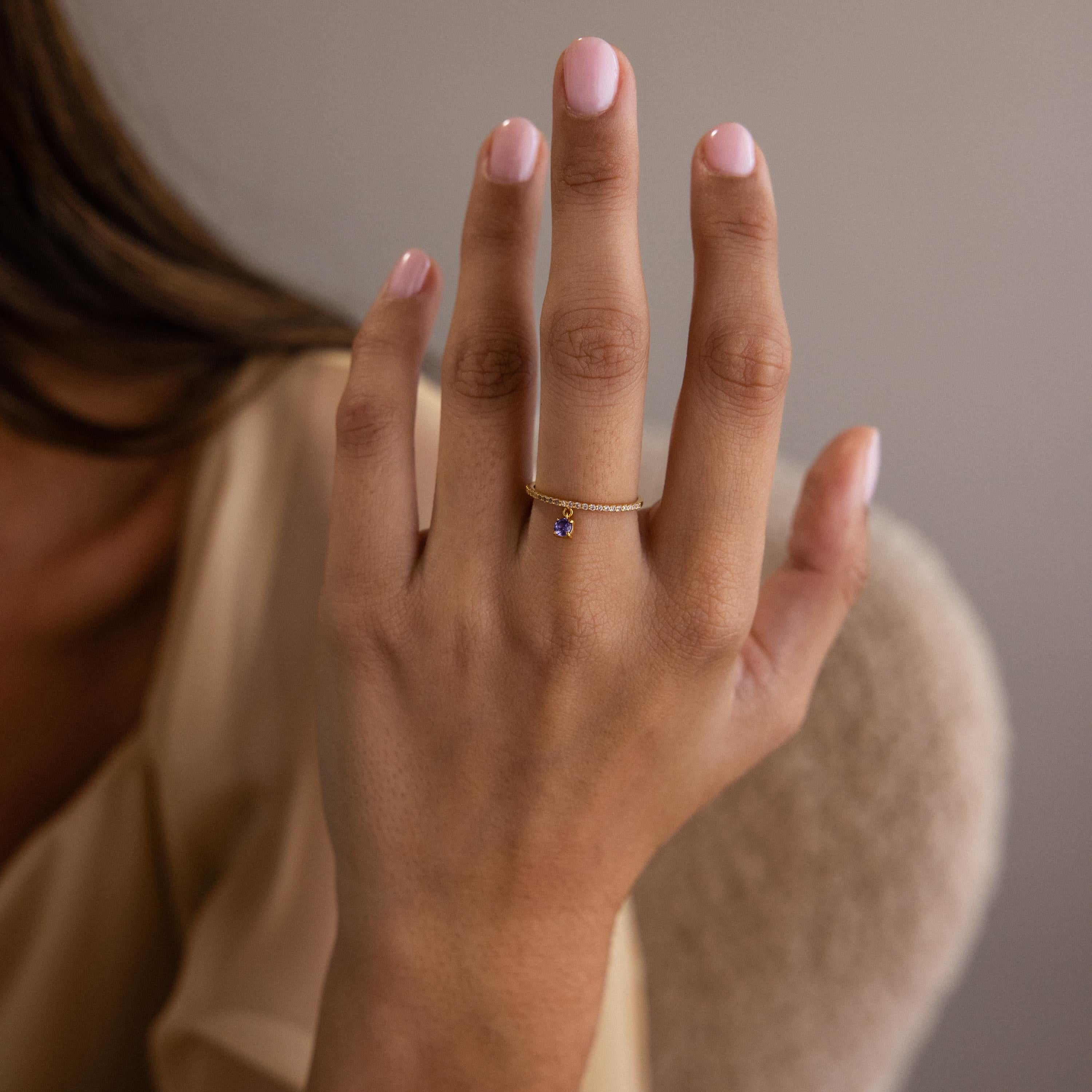 A hand with light pink nails wears the delicate gold Pave Birthstone Drop Ring featuring a small purple gemstone charm.