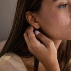 A woman touches her ear while wearing the Pave Birthstone Drop Ring, its gold and gemstone details beautifully highlighted by soft lighting on her face.