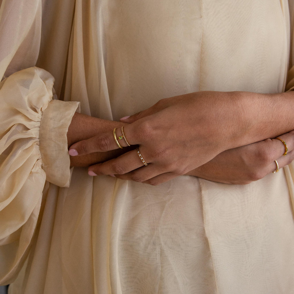 Close-up of crossed hands wearing gold rings, featuring the Pave Birthstone Drop Ring with delicate diamond accents, set against sheer beige, long-sleeved flowy fabric in the background.