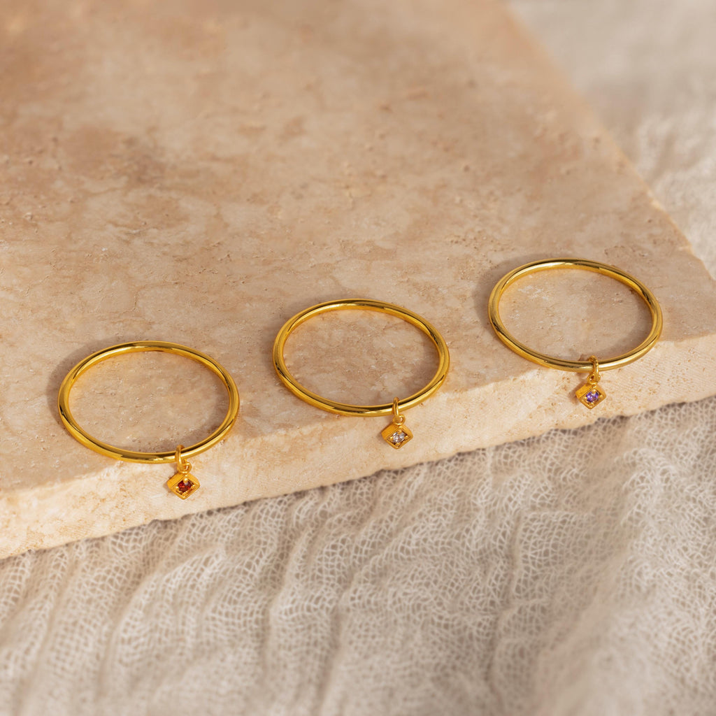 Three Dainty Birthstone Drop Rings, each featuring a small colored gemstone charm, are displayed on a beige textured stone surface—ideal for anyone seeking personalized, meaningful jewelry with a pop of color.