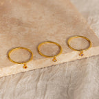 Three Dainty Birthstone Drop Rings, each featuring a small colored gemstone charm, are displayed on a beige textured stone surface—ideal for anyone seeking personalized, meaningful jewelry with a pop of color.