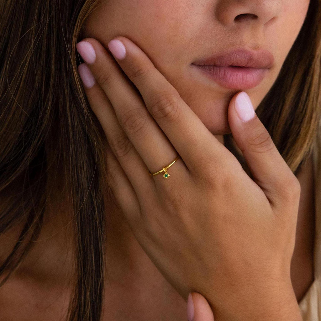 A woman with natural nails wears the Dainty Birthstone Drop Ring, a thin gold band featuring a delicate birthstone drop—an elegant, personalized jewelry piece as she touches her face.