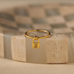 The Initial Padlock Drop Ring features a small lock charm engraved with the letter B, displayed on a beige patterned surface—a stylish, personalized jewelry piece.