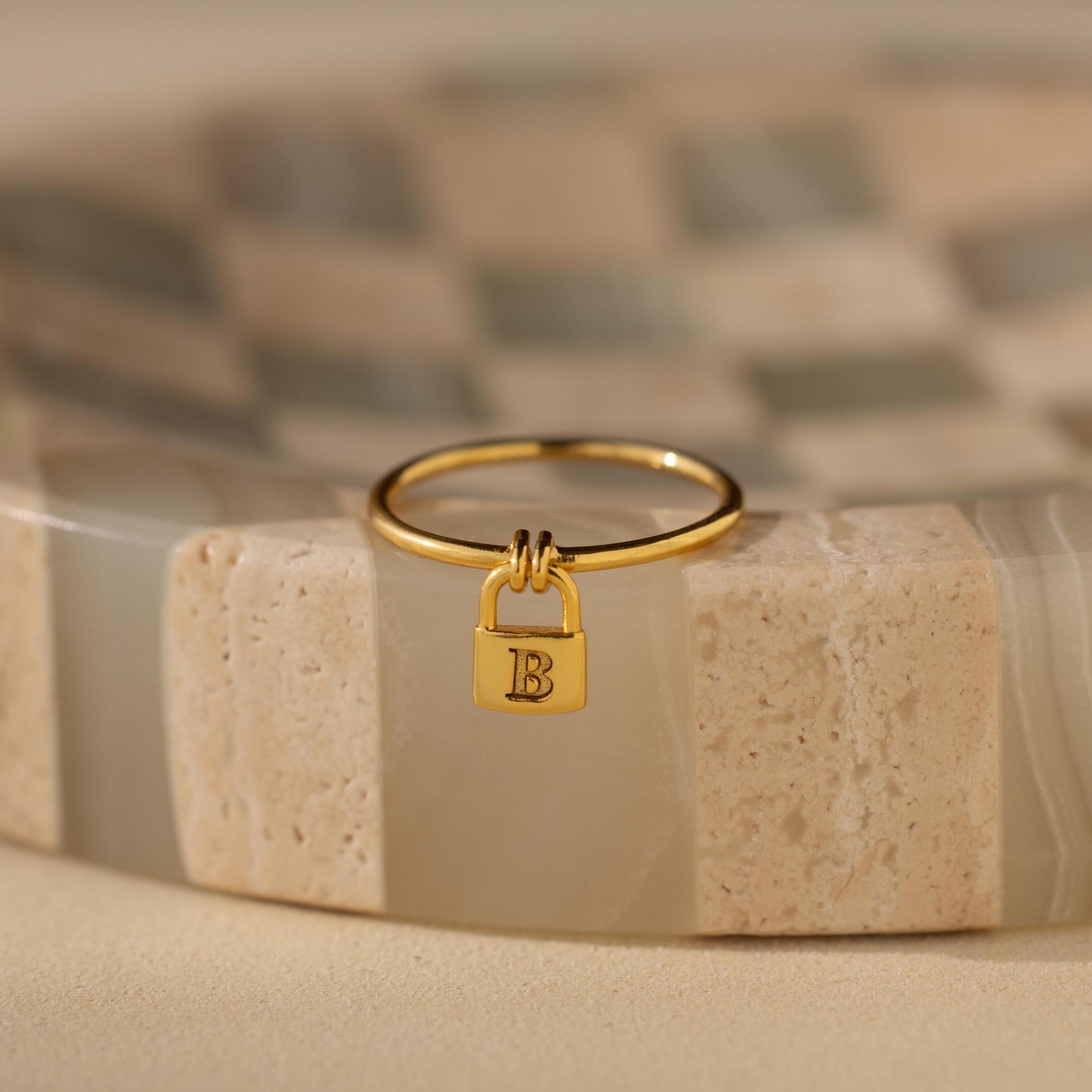 The Initial Padlock Drop Ring features a small lock charm engraved with the letter B, displayed on a beige patterned surface—a stylish, personalized jewelry piece.