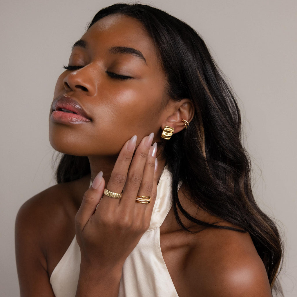 A woman with long hair and closed eyes, wearing a white halter top, poses with her hand on her face while showcasing Bold Square Wave Studs luxury earrings.