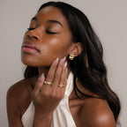 A woman with long hair and closed eyes, wearing a white halter top, poses with her hand on her face while showcasing Bold Square Wave Studs luxury earrings.