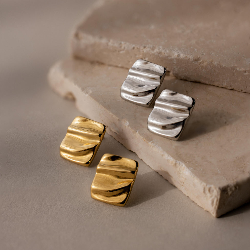 Two pairs of Bold Square Wave Studs in gold and silver are displayed on textured beige stone tiles, perfect for those who love minimalist jewelry.