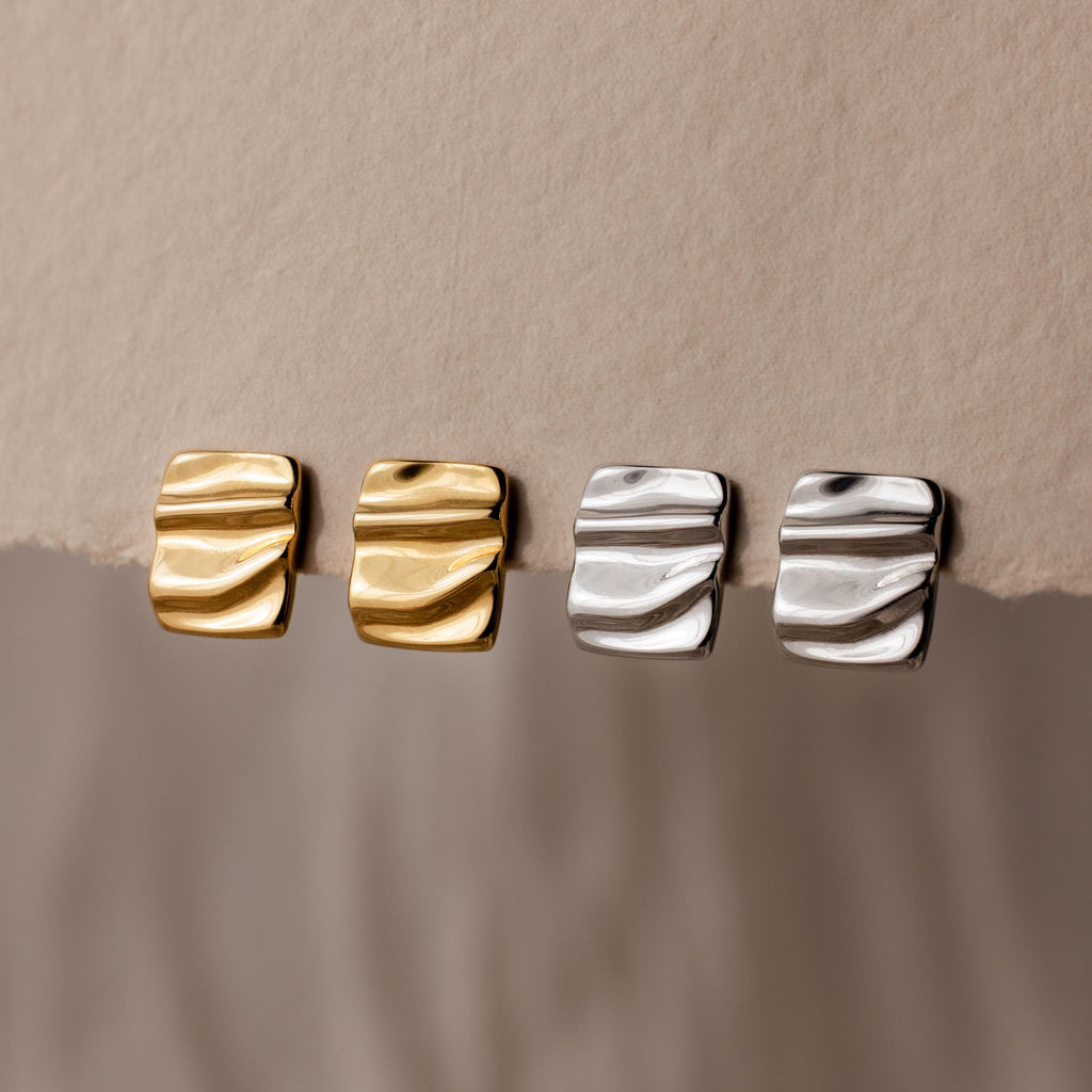 Two pairs of Bold Square Wave Studs, one in gold and one in silver, displayed on textured beige paper—a refined touch for any luxury jewelry lover.