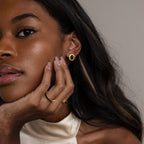 A woman, adorned in gold jewelry and Vintage Oval Black Studs, rests her chin on her hand against a neutral background.