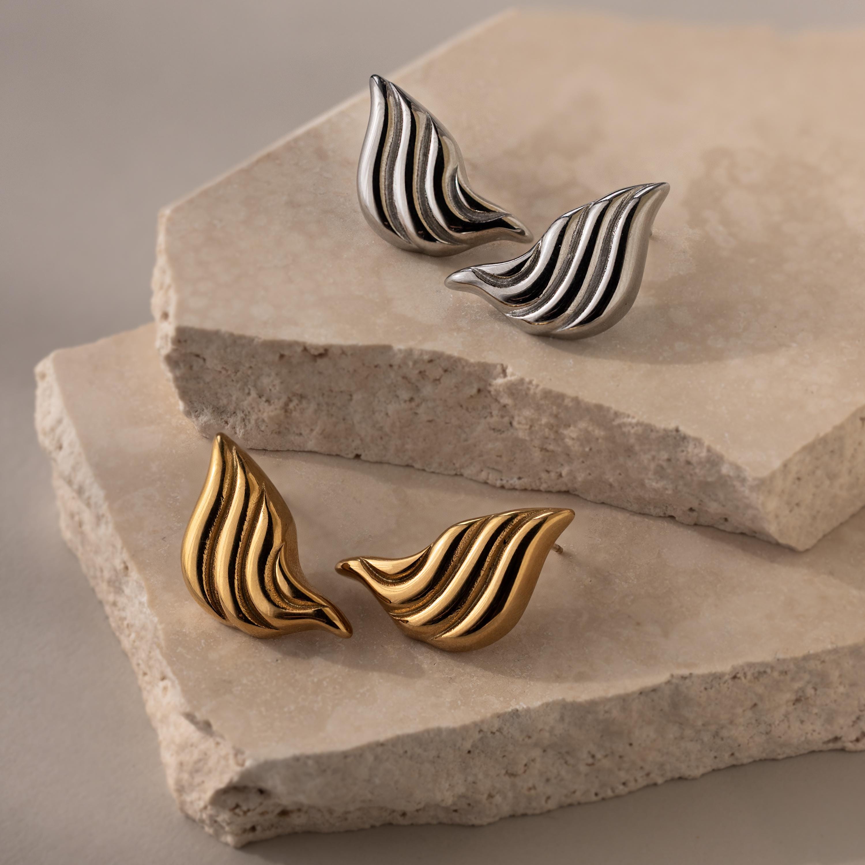 Bold Wing Studs in gold and silver with an abstract, wave-shaped wing design are displayed on stacked stone slabs.