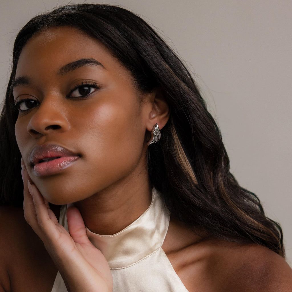 Woman with long dark hair wearing a cream halter top and Bold Wing Studs, gently touching her face.