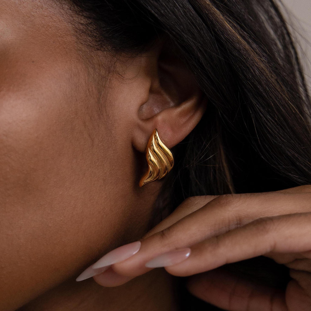 Close-up of Bold Wing Studs, gold abstract wing earrings, worn on an ear with a hand near the face and nude-painted nails.