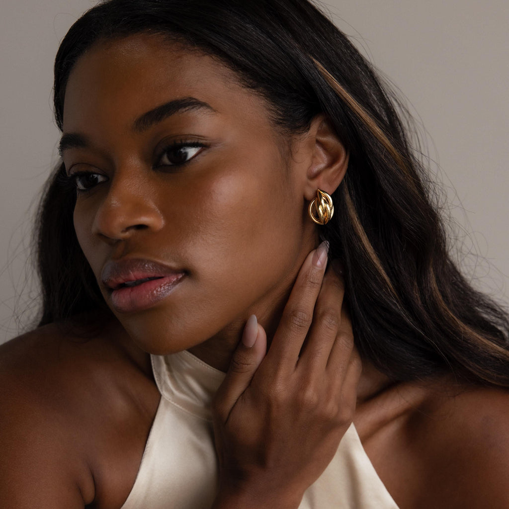 A woman with long hair wears a cream halter top and Bold Multi Knot Studs, touching her neck thoughtfully and embodying modern elegance.