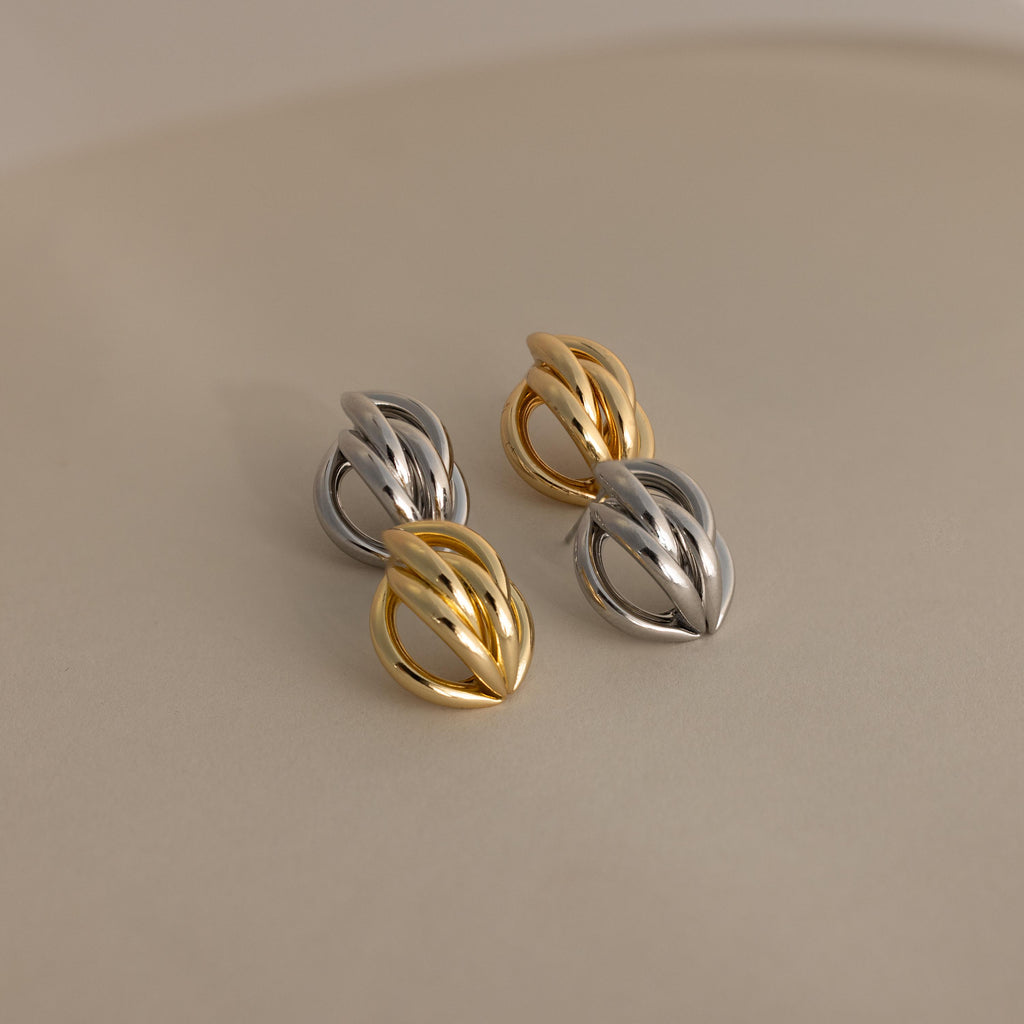 The Bold Multi Knot Studs showcase intertwined gold and silver in a modern knot design, exuding elegance, and are displayed on a smooth, neutral surface.