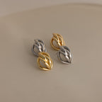 The Bold Multi Knot Studs showcase intertwined gold and silver in a modern knot design, exuding elegance, and are displayed on a smooth, neutral surface.