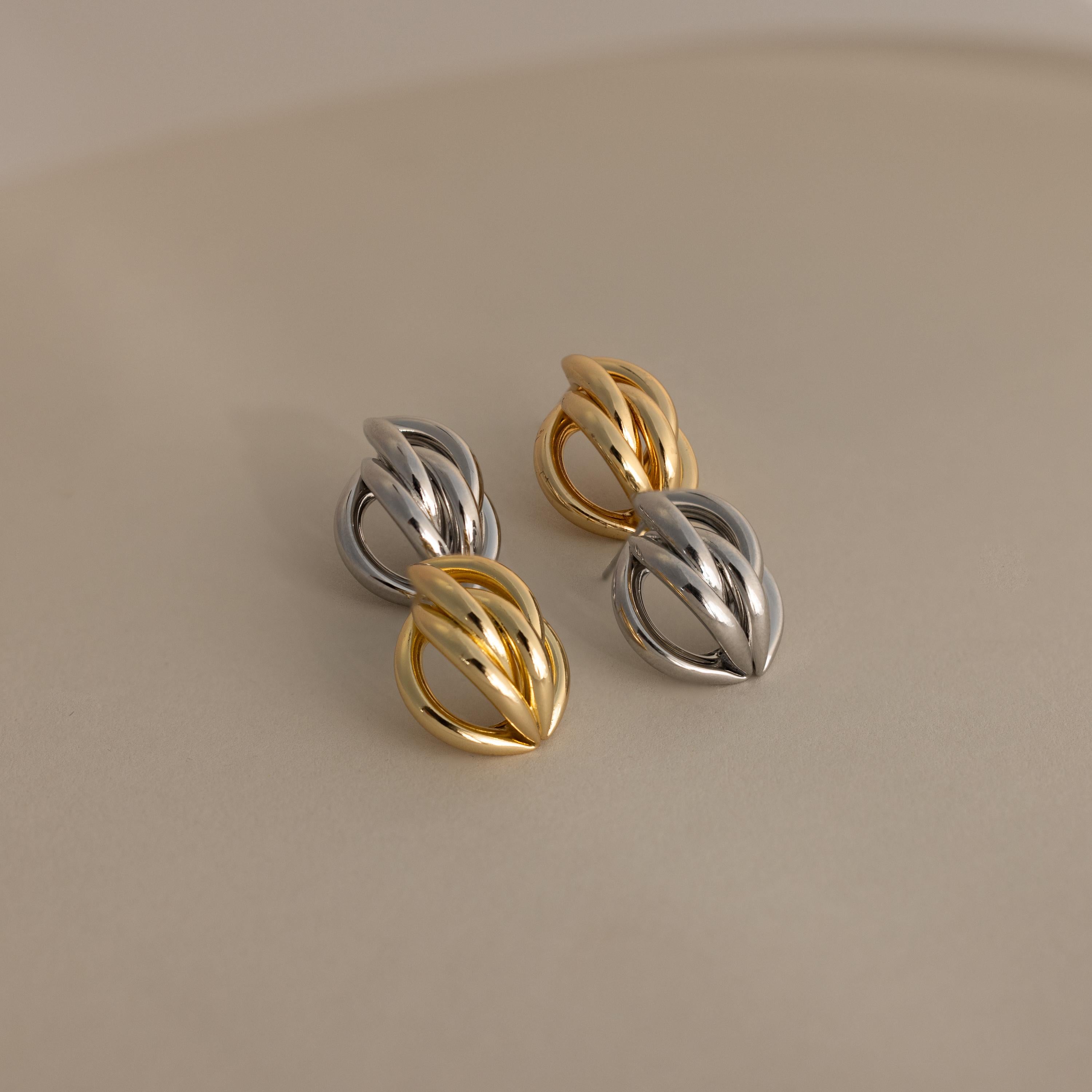The Bold Multi Knot Studs showcase intertwined gold and silver in a modern knot design, exuding elegance, and are displayed on a smooth, neutral surface.
