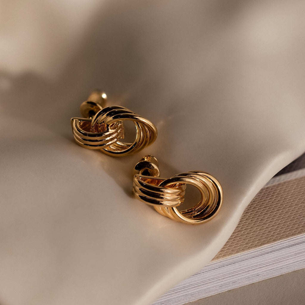 Micro Triple Knot Earrings rest on a soft, beige textured fabric—an elegant nod to quiet luxury.