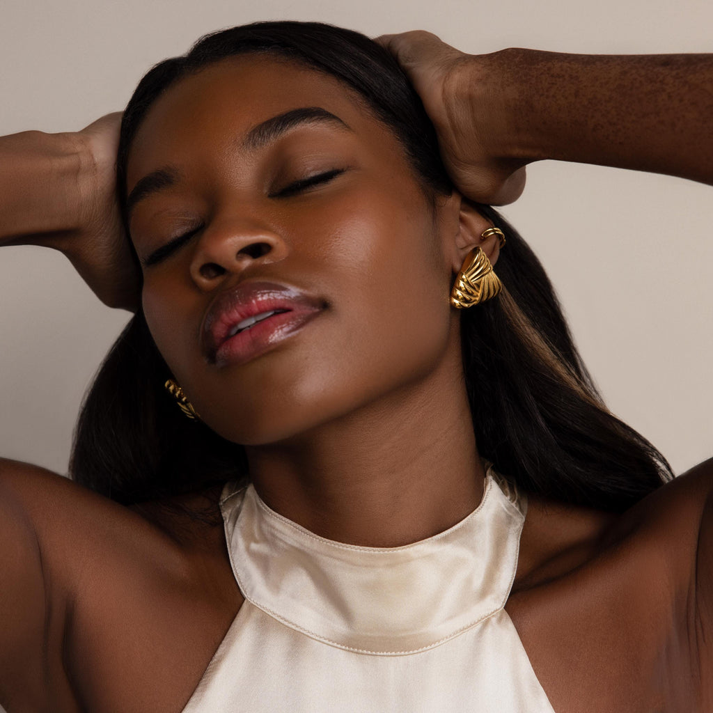 With her eyes closed and hands resting on her head, a woman in a cream satin halter top showcases the Bold Triangle Knot Studs—her ultimate statement accessory.