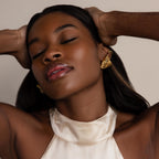 With her eyes closed and hands resting on her head, a woman in a cream satin halter top showcases the Bold Triangle Knot Studs—her ultimate statement accessory.
