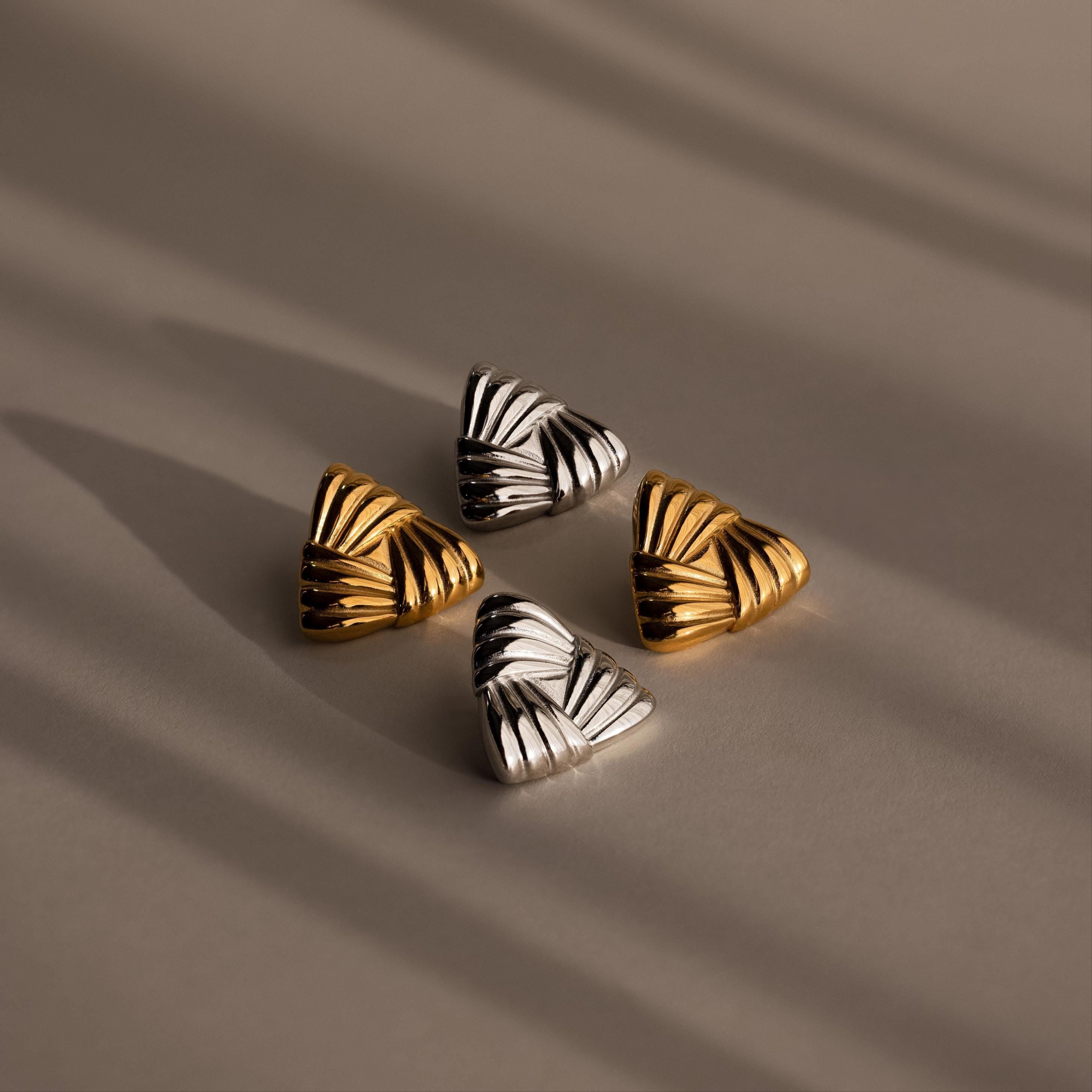 The Bold Triangle Knot Studs—two gold and two silver triangular earrings with engraved lines—are arranged on a neutral, softly lit surface, creating a statement look ideal for lovers of chunky earrings.
