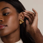 A woman with natural makeup and a neutral background touches her ear, showcasing the Bold Triangle Knot Studs—chunky earrings that make a striking statement.
