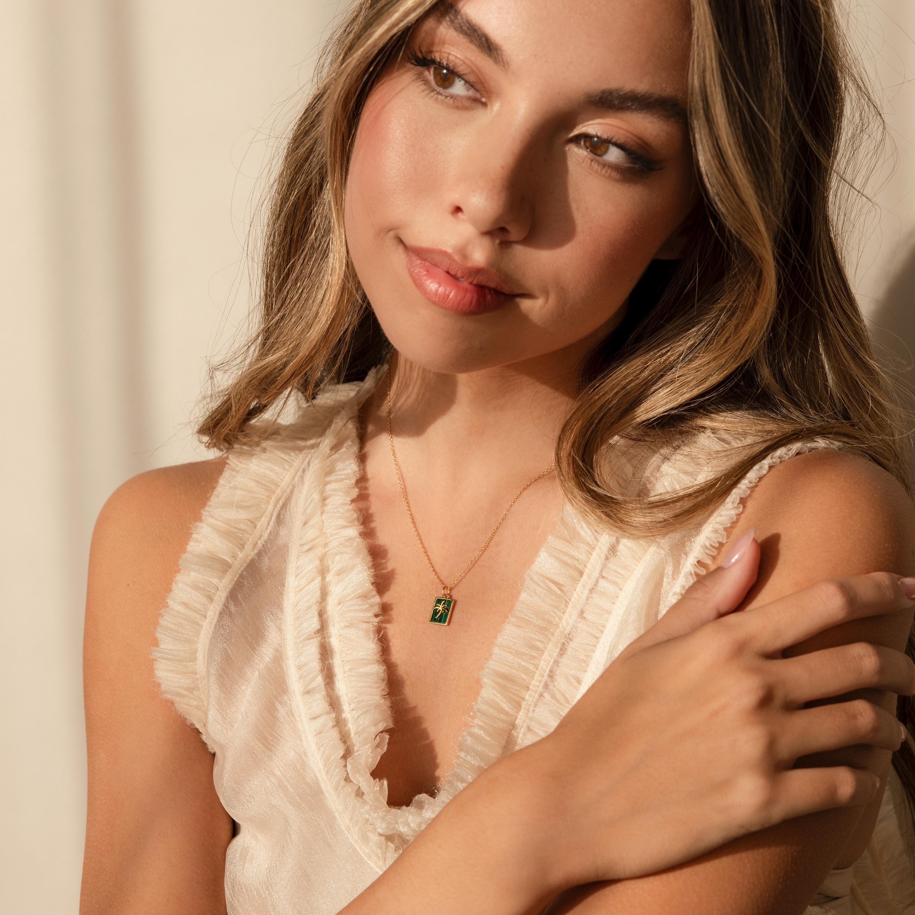 Woman with long hair wears a sleeveless ruffled cream top and the Malachite Palm Tree Necklace, featuring a delicate malachite gemstone, as she gazes thoughtfully to the side.